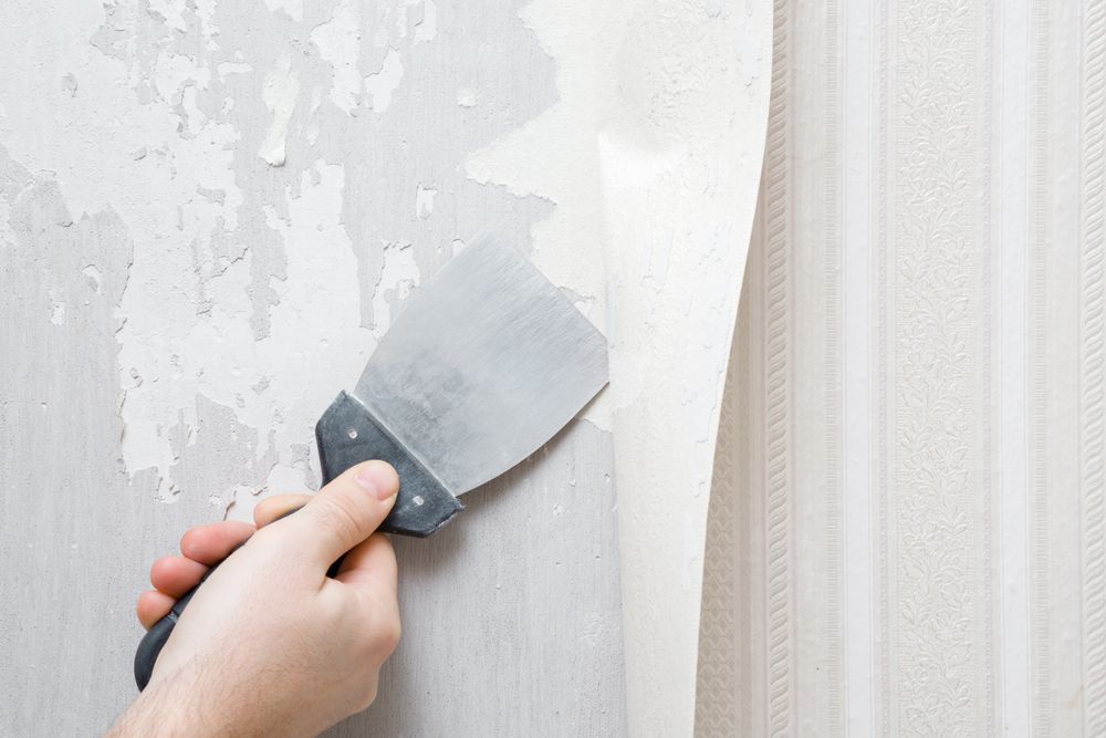 Hand using a putty knife to scrape peeling paint from a white wall