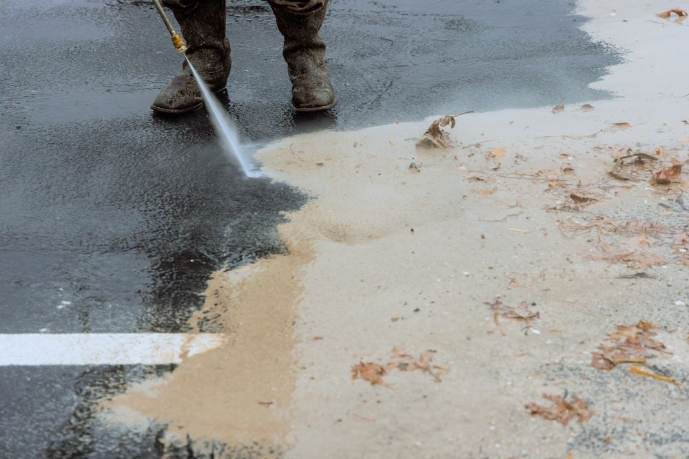 Worker pressure-washing a wet parking lot, clearing dirt from a painted curb line