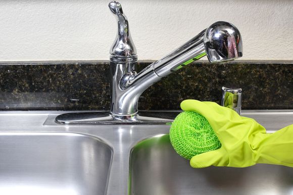 A person wearing yellow gloves is cleaning a sink with a green sponge