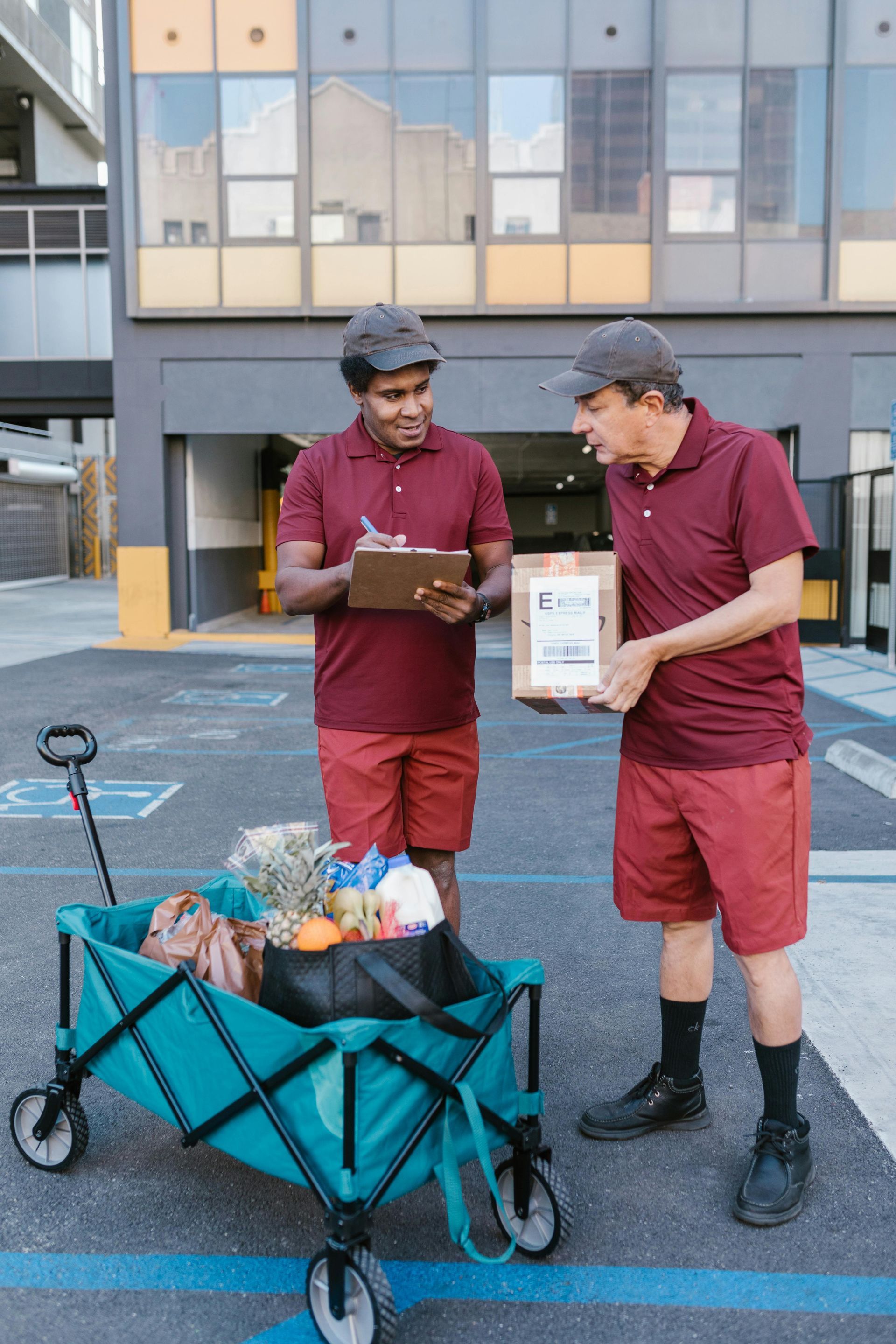 Two delivery men are standing next to a wagon filled with groceries.