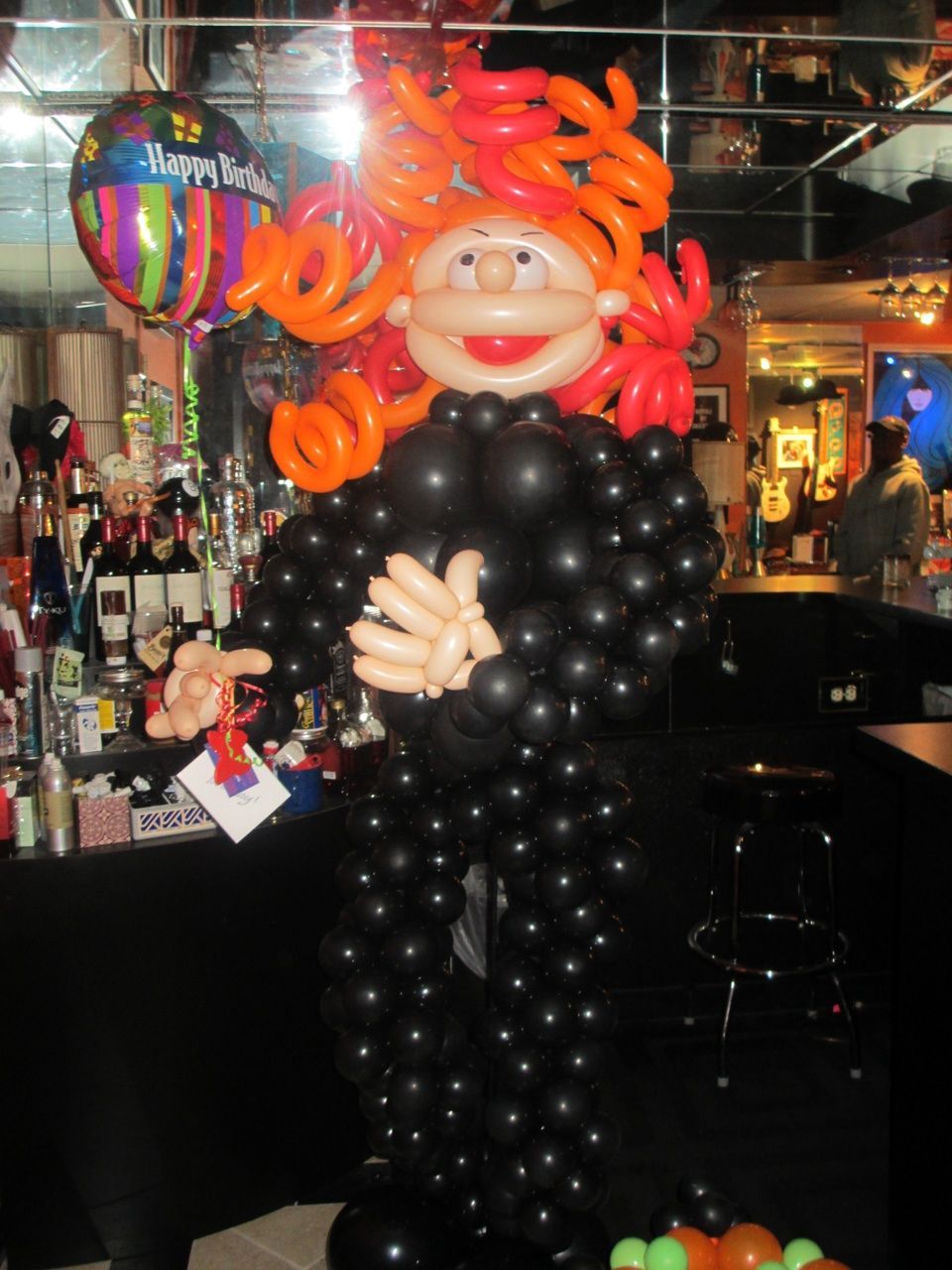 A balloon sculpture of a woman with red hair is in front of a happy birthday balloon