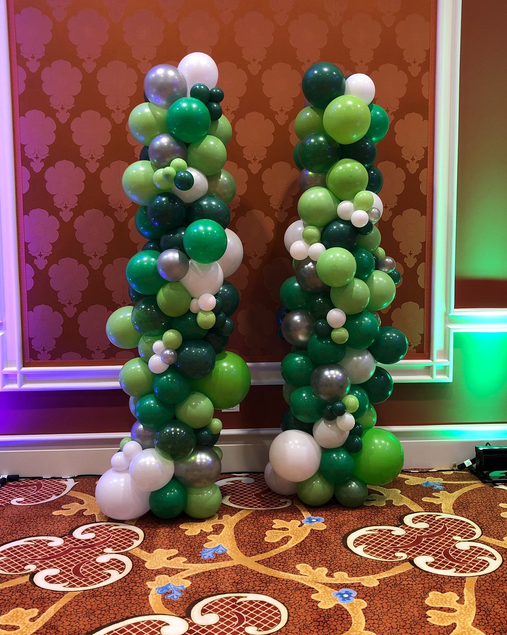 Two columns made of green , white , and silver balloons are sitting on a carpet.
