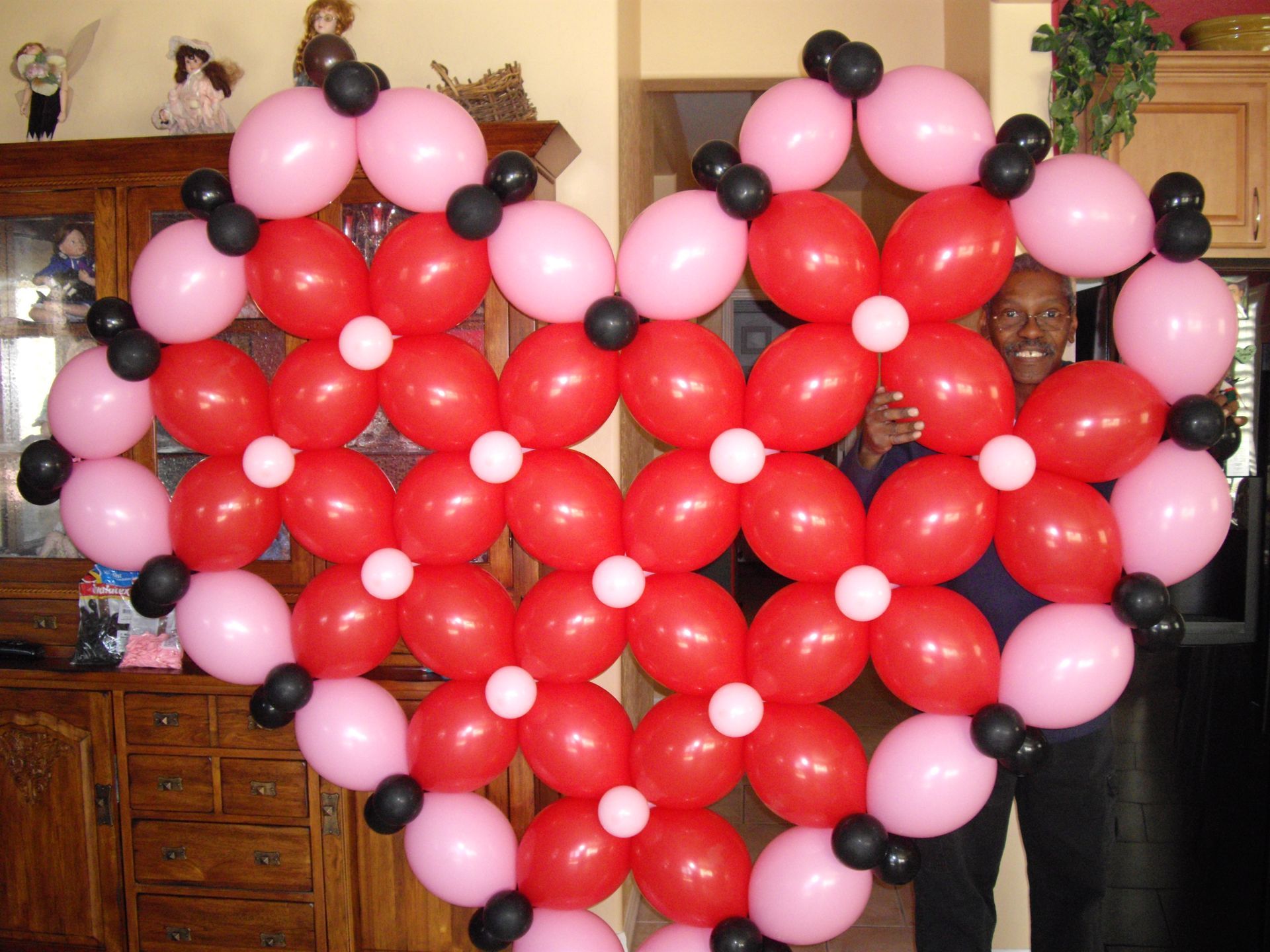 A man stands in front of a heart made of red and pink balloons