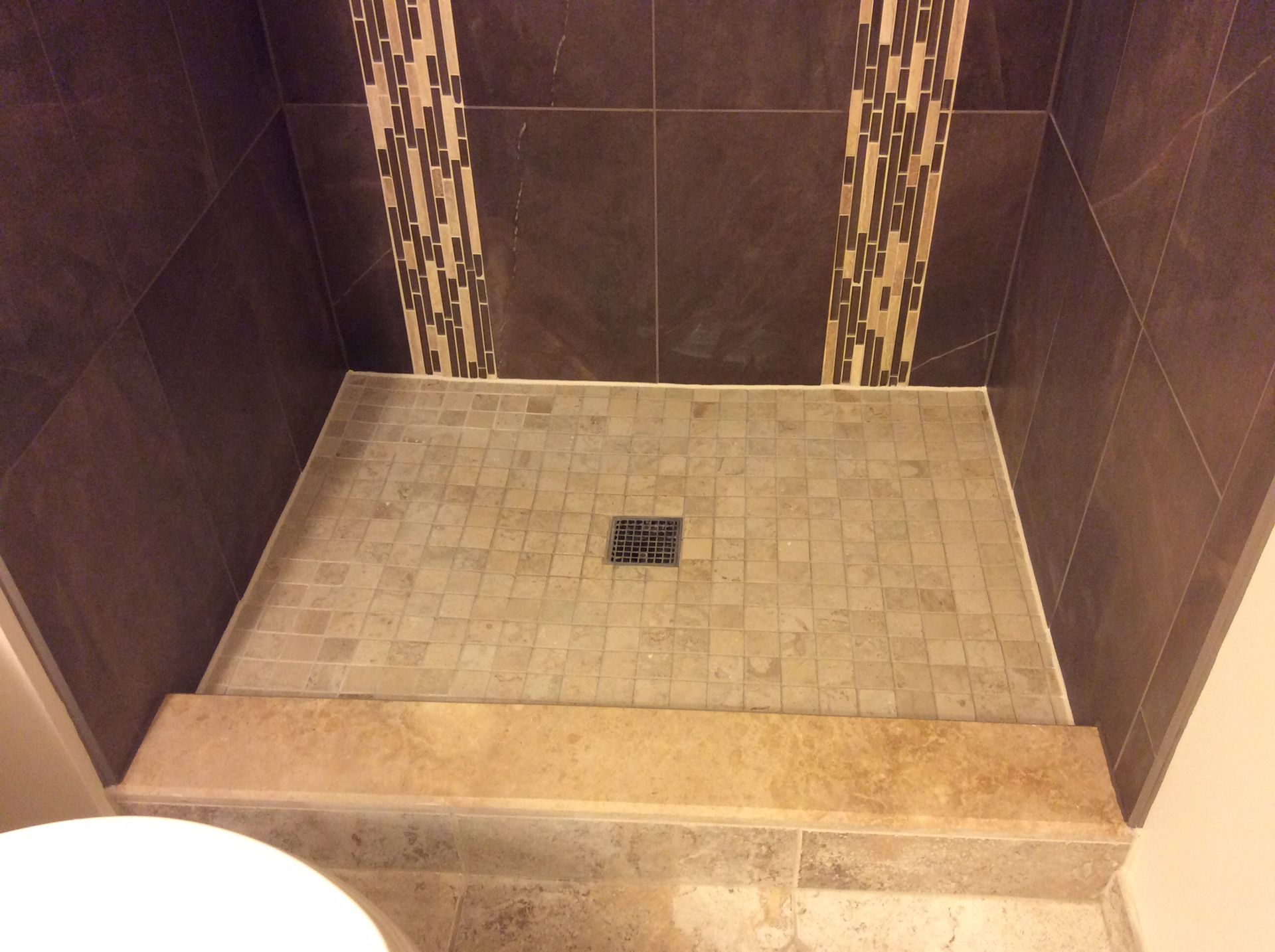 Shower stall with brown and tan tiles, mosaic shower floor, and a small curb.