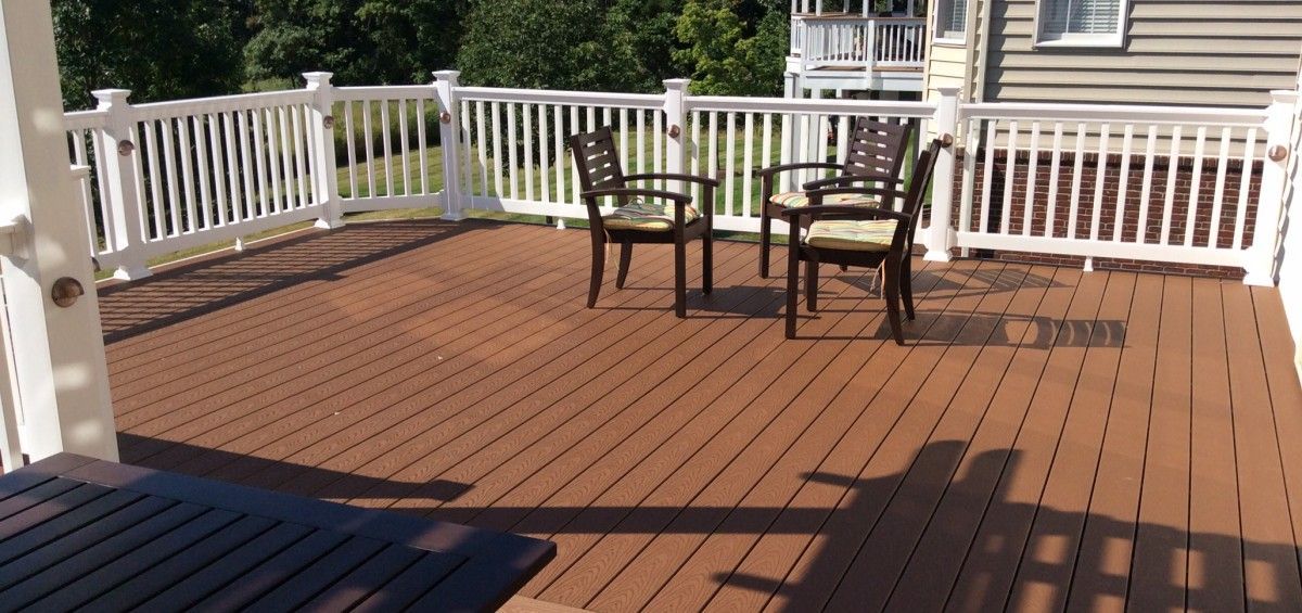 Wooden deck with white railing, three chairs, and a house in the background on a sunny day.