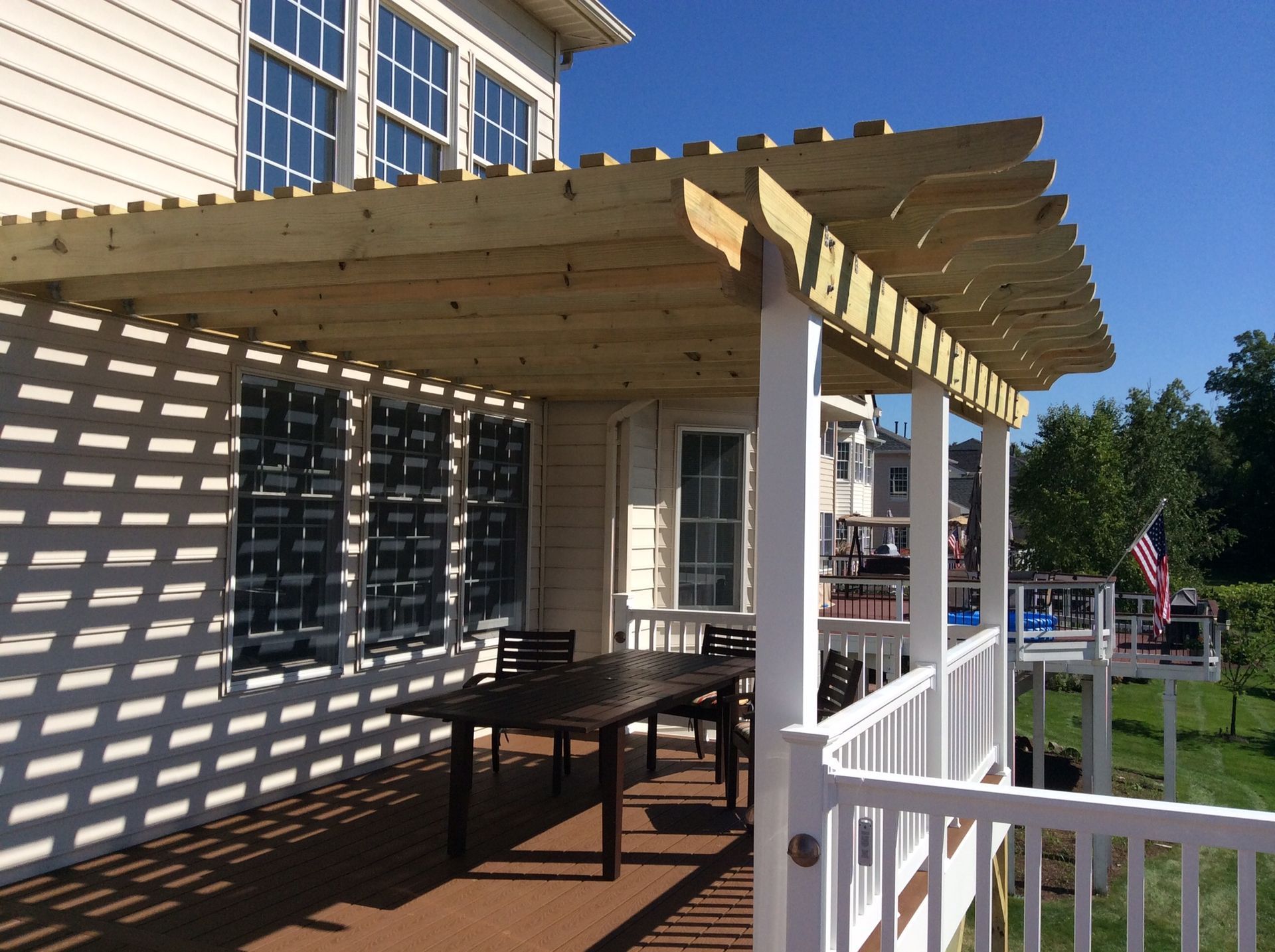 Wooden pergola over a deck with a table and chairs, attached to a house with an American flag.