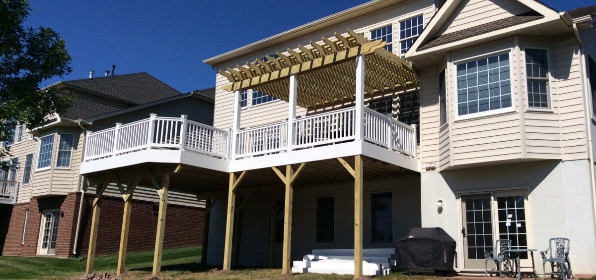 Multi-level deck with white railing and pergola, attached to a light-colored house under a clear blue sky.