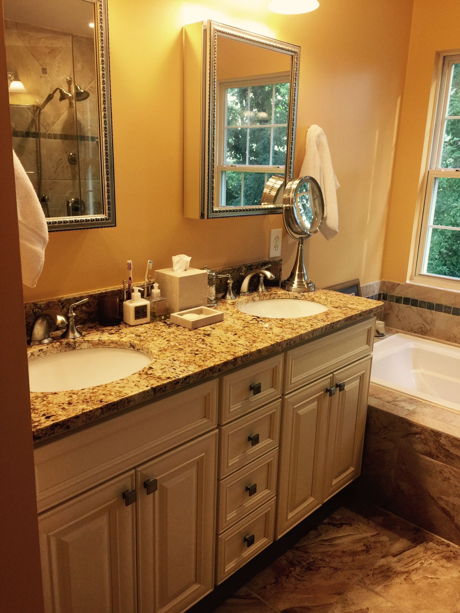 Bathroom with double sinks, light granite countertop, cream cabinets, and a bathtub near a window.
