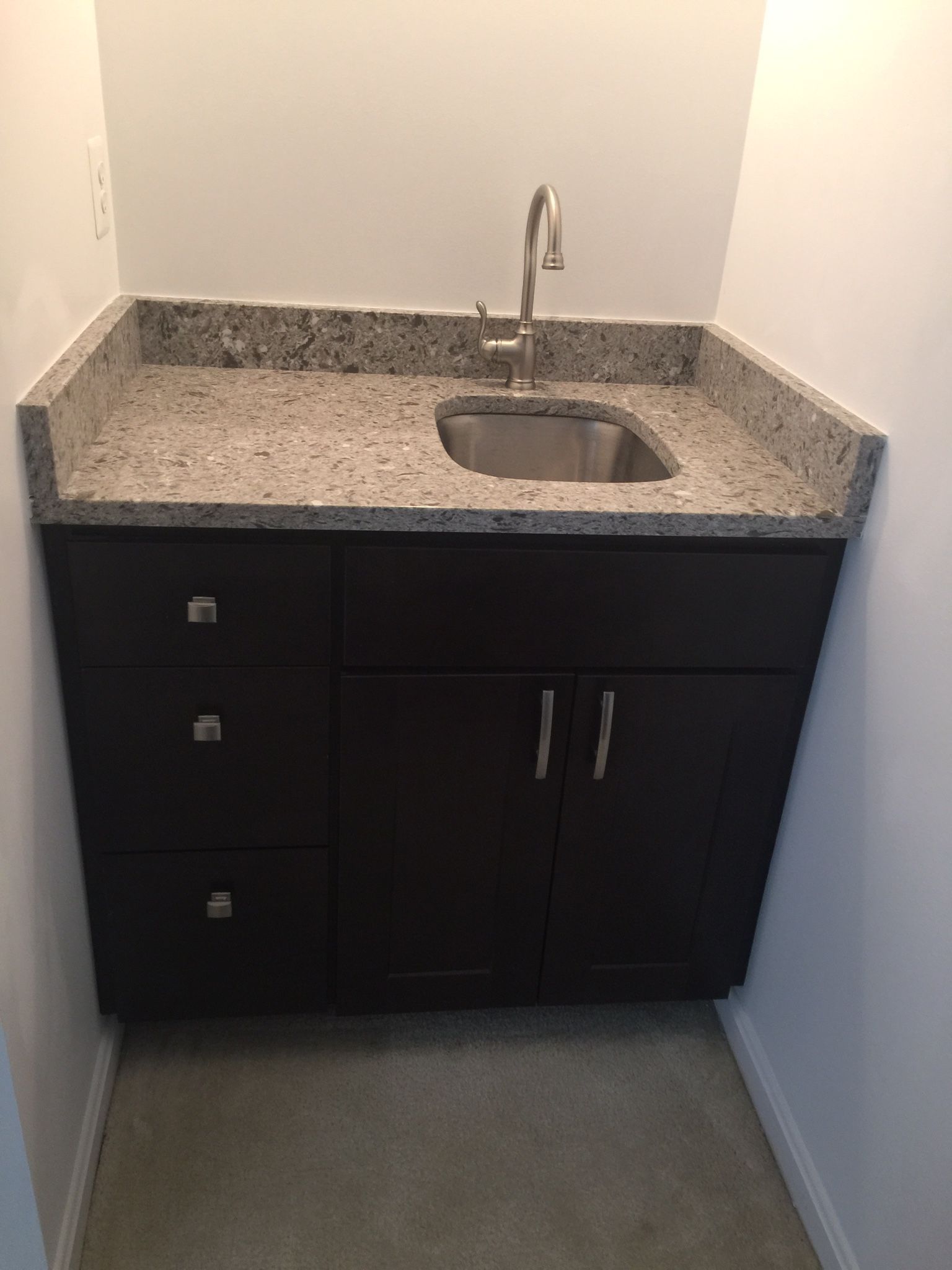 Dark brown sink cabinet with granite countertop and stainless steel sink. Silver faucet. Light gray walls.