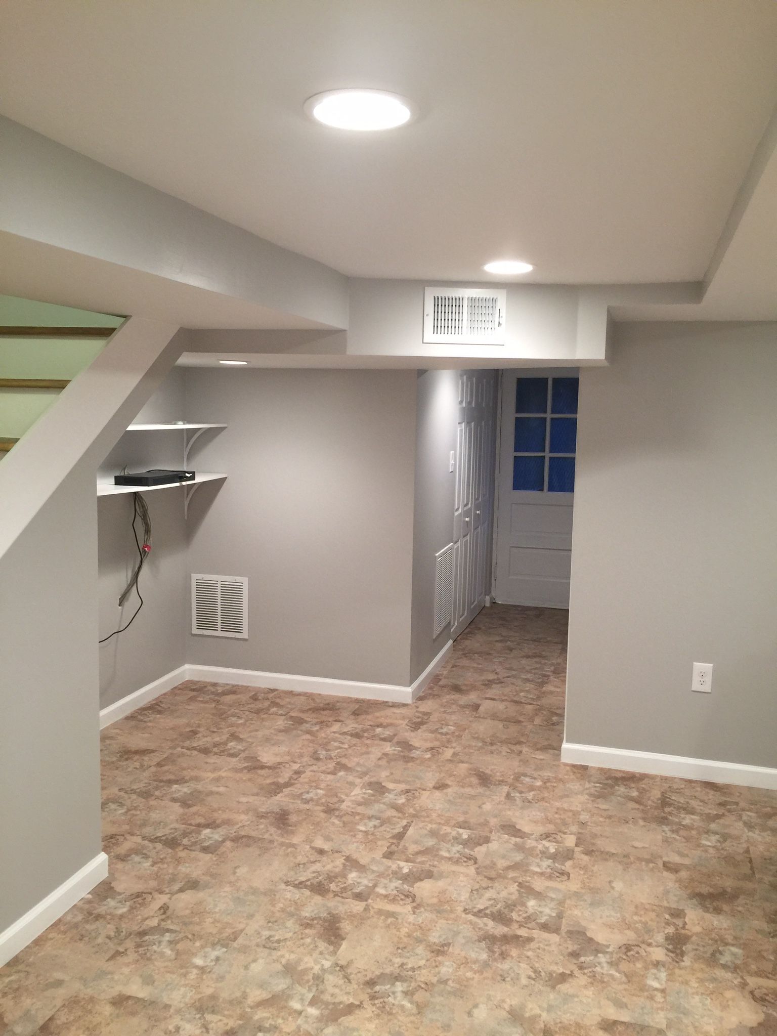 Gray basement with tiled floor, white trim, recessed lights, shelves, and a doorway.