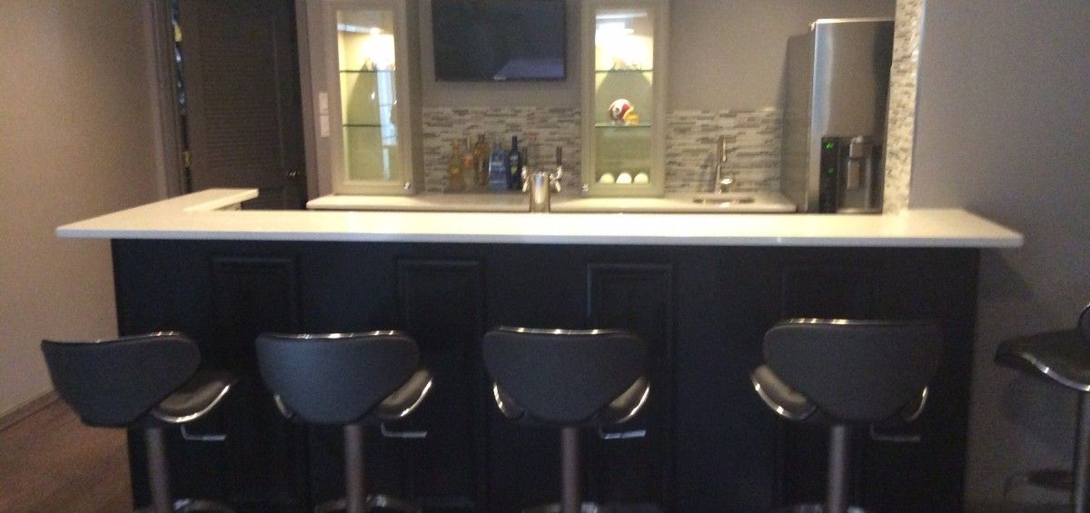 A home bar with black stools, dark cabinets, a white countertop, and a built-in backsplash.