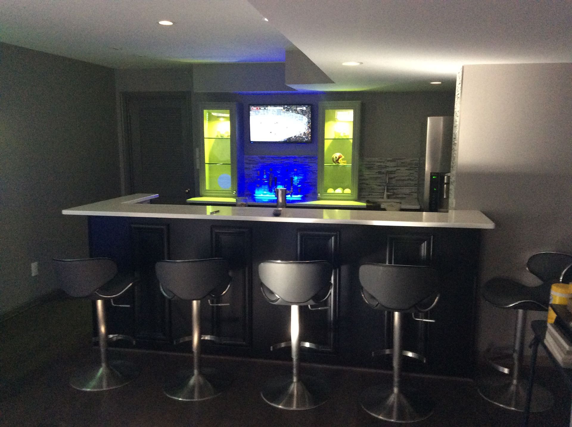 Basement bar with dark cabinets, white countertop, and black stools. TV and illuminated shelves in the background.