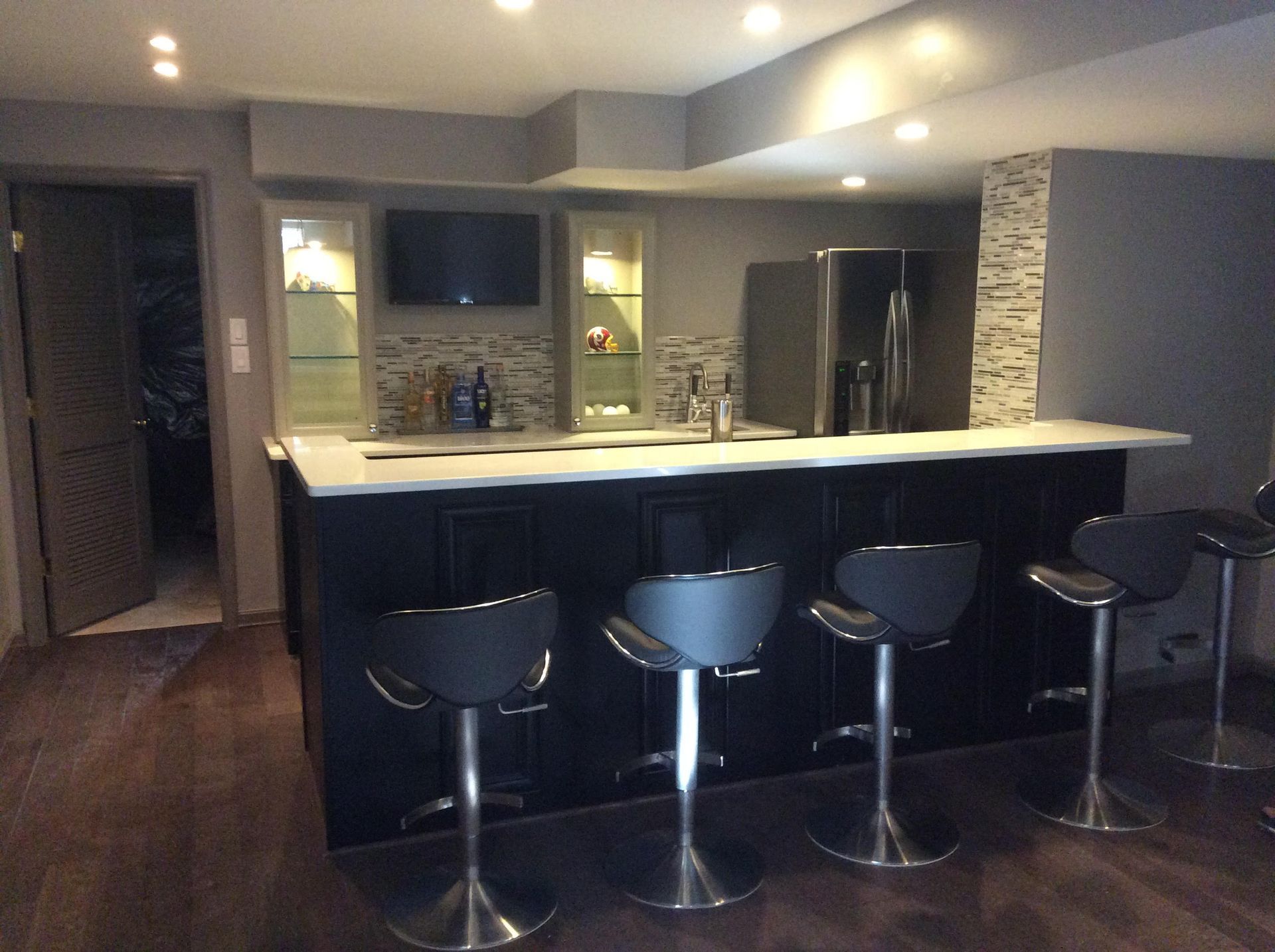 Basement bar with black cabinets, white countertop, and black bar stools. Grey walls with a fridge and TV.