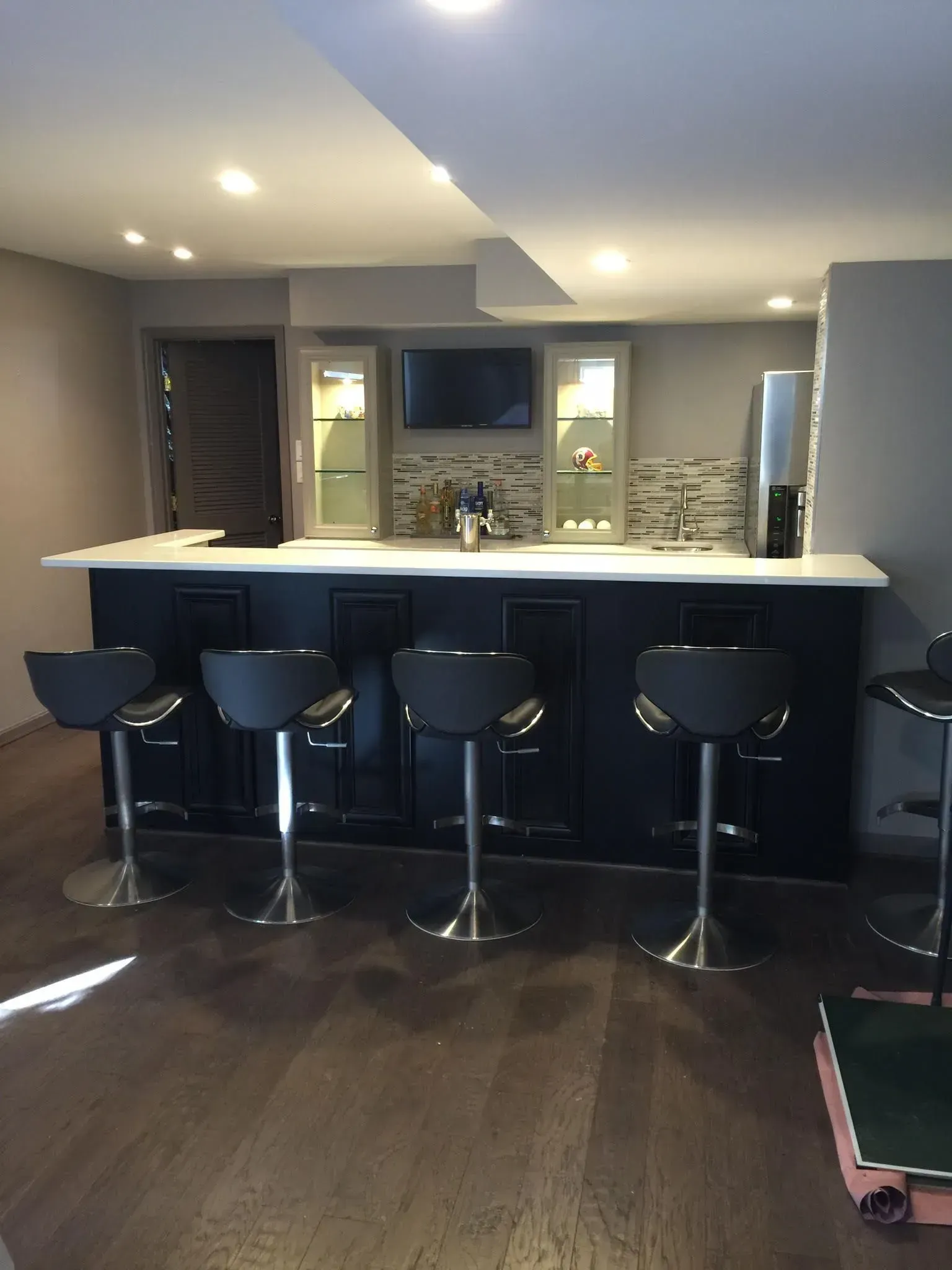 Basement bar with black cabinets, white countertop, and gray walls. Bar stools line the counter.