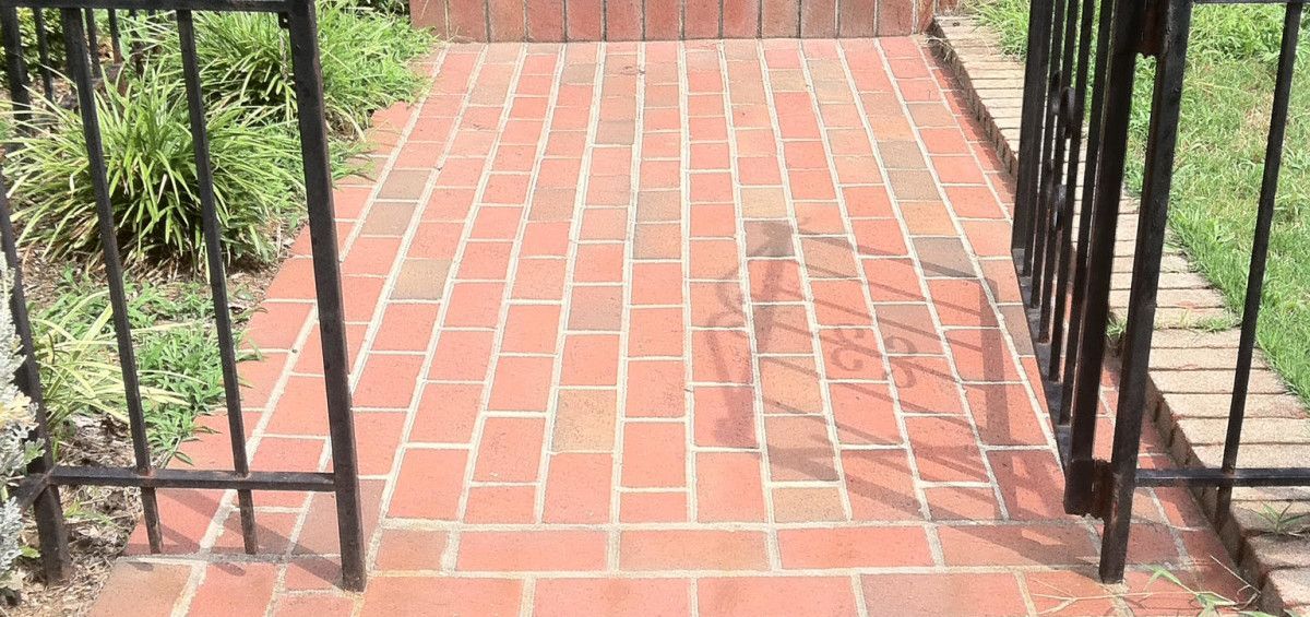 Brick pathway leads from wrought iron gate towards a brick wall, with green plants on either side.
