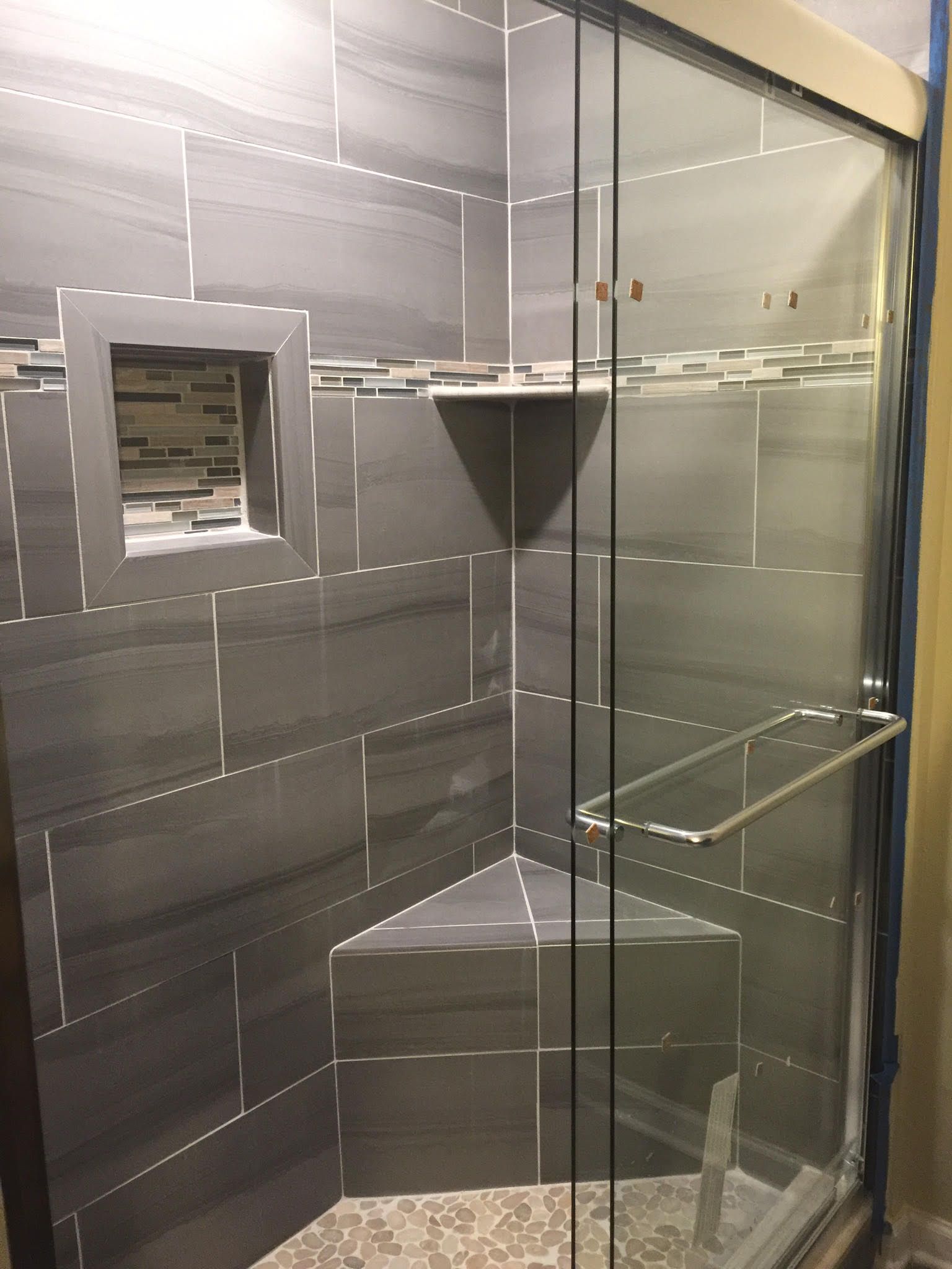 Gray tiled shower with glass door and built-in corner seat. Features a niche and a towel bar.