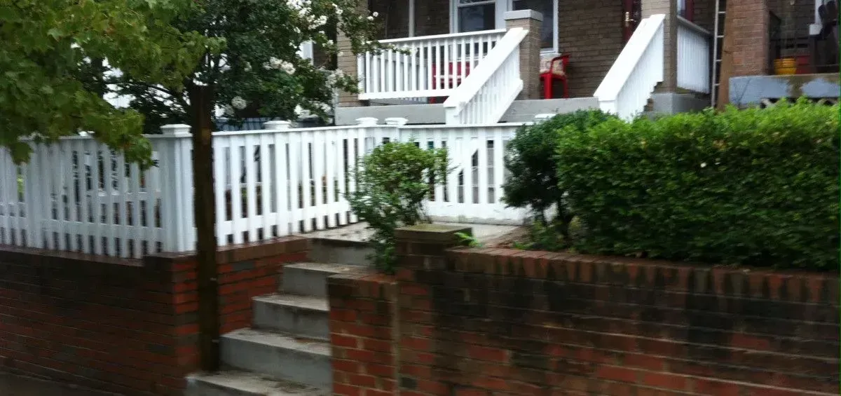 White picket fence and steps leading up to a house with a porch, surrounded by greenery.