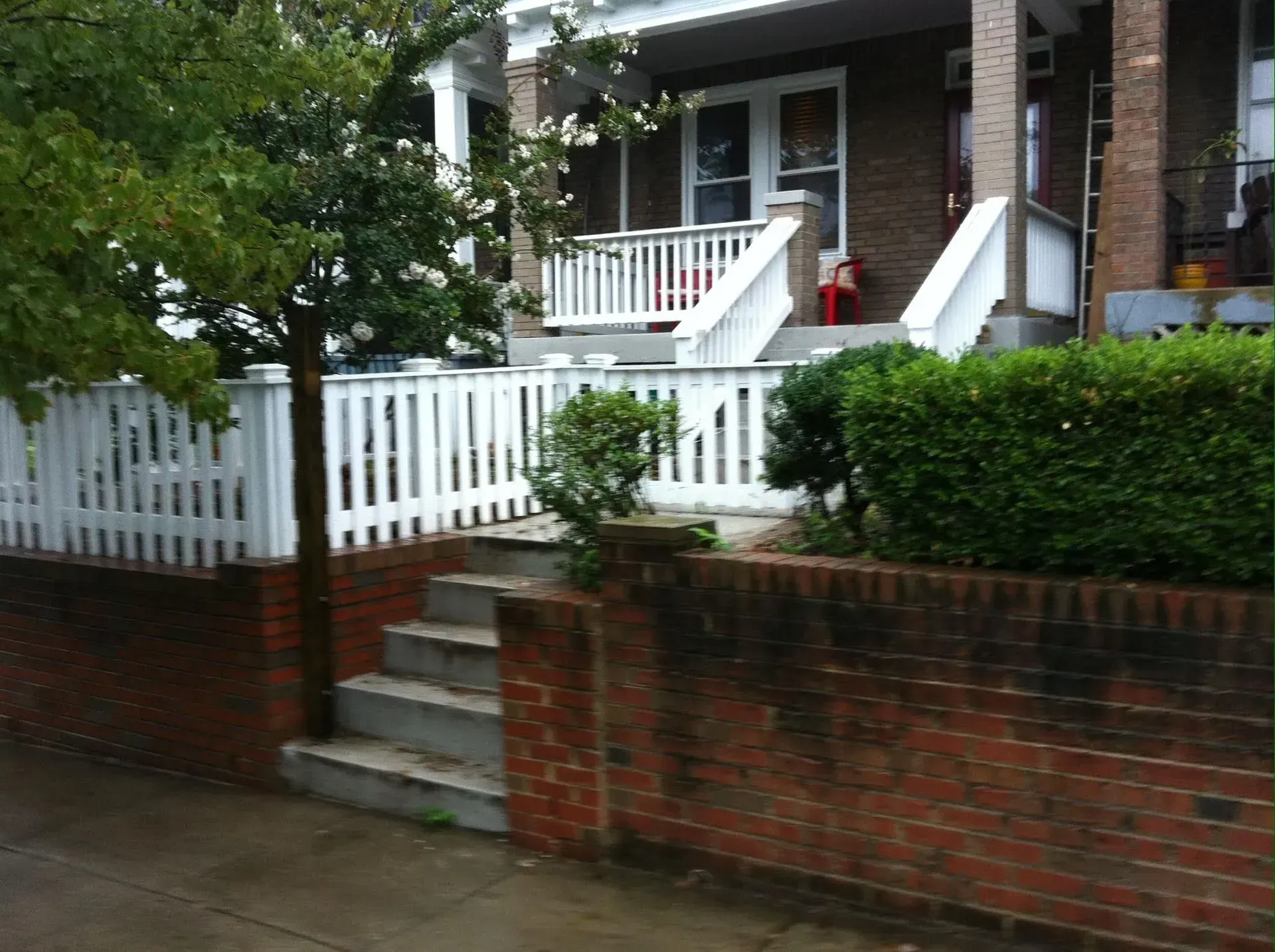 Brick house with white picket fence, stairs, and green shrubs.