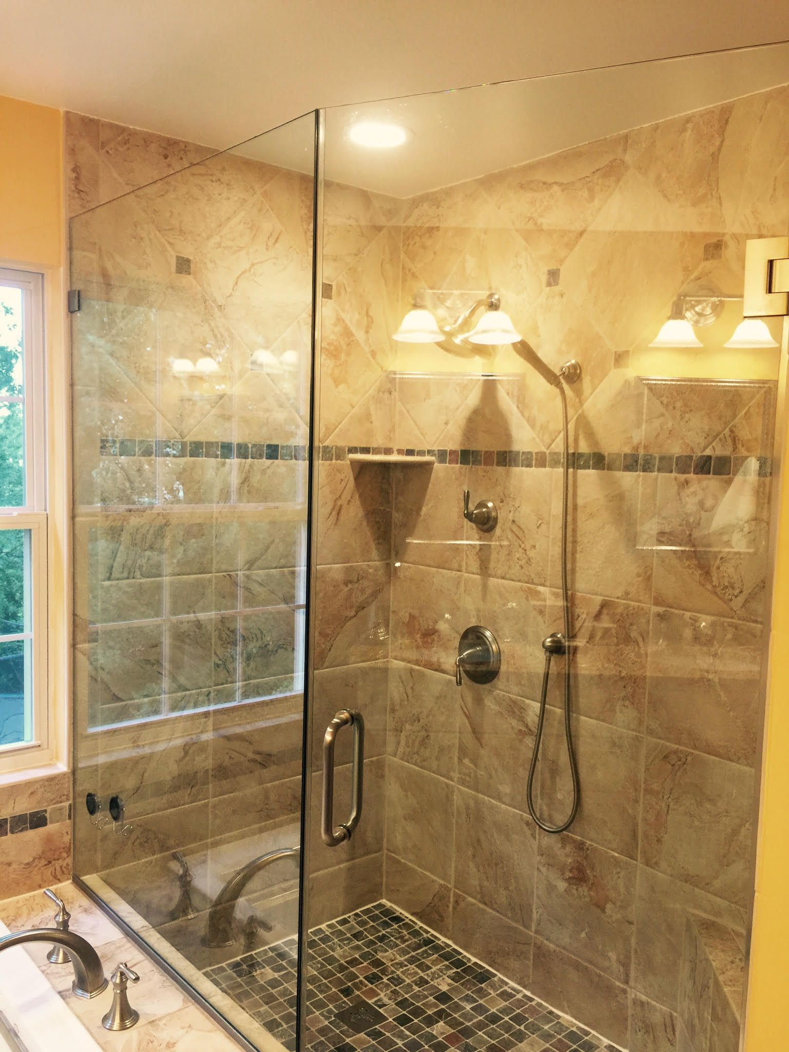 Shower with glass doors and beige tile walls. Features include a showerhead, handle, and built-in shelving.
