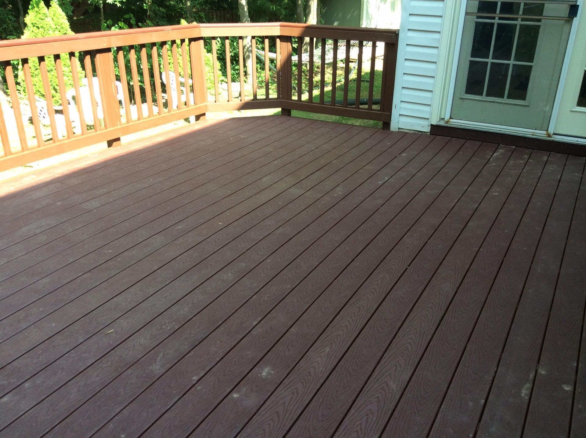 Wooden deck with brown planks and railing. Sunlight, next to white siding and door.