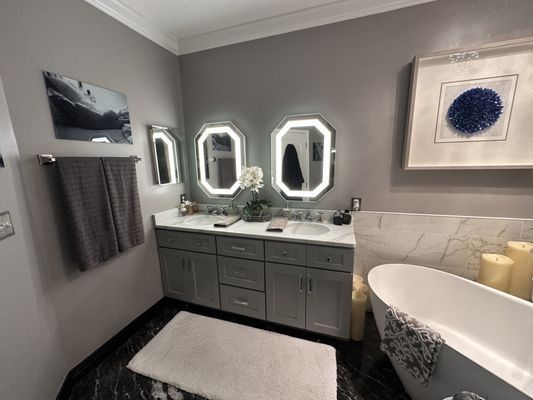 Gray and white bathroom with double vanity, lighted mirrors, and a freestanding tub.