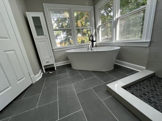 Bathroom with a free-standing tub by windows, grey tile floor, and a white cabinet.