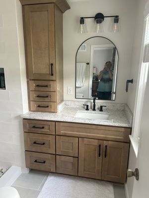 Bathroom with light wood vanity, arched mirror, black fixtures, and a tall cabinet.