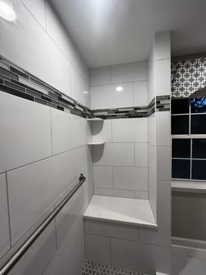 Modern, accessible shower with white tile, a built-in bench, shelves, and a stainless steel grab bar.