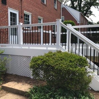 White deck with black railing, lattice base, and stairs, in front of a brick building.