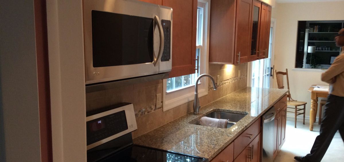 A kitchen with brown cabinets, granite countertops, and stainless steel appliances. A person walks by.
