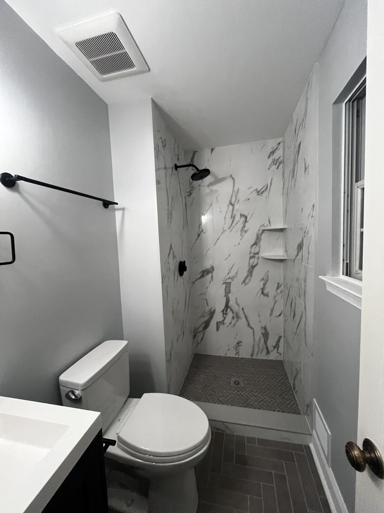 A small bathroom with a marble-tiled shower, toilet, sink, and gray walls.