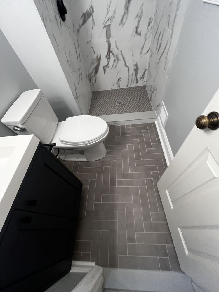Small bathroom with gray herringbone floor tile, white toilet, black vanity, marble shower, and open white door.