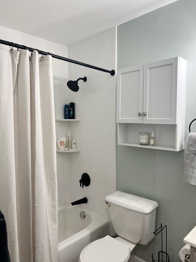 White bathroom with bathtub, toilet, and storage cabinet. Pale green wall, black fixtures, and white shower curtain.