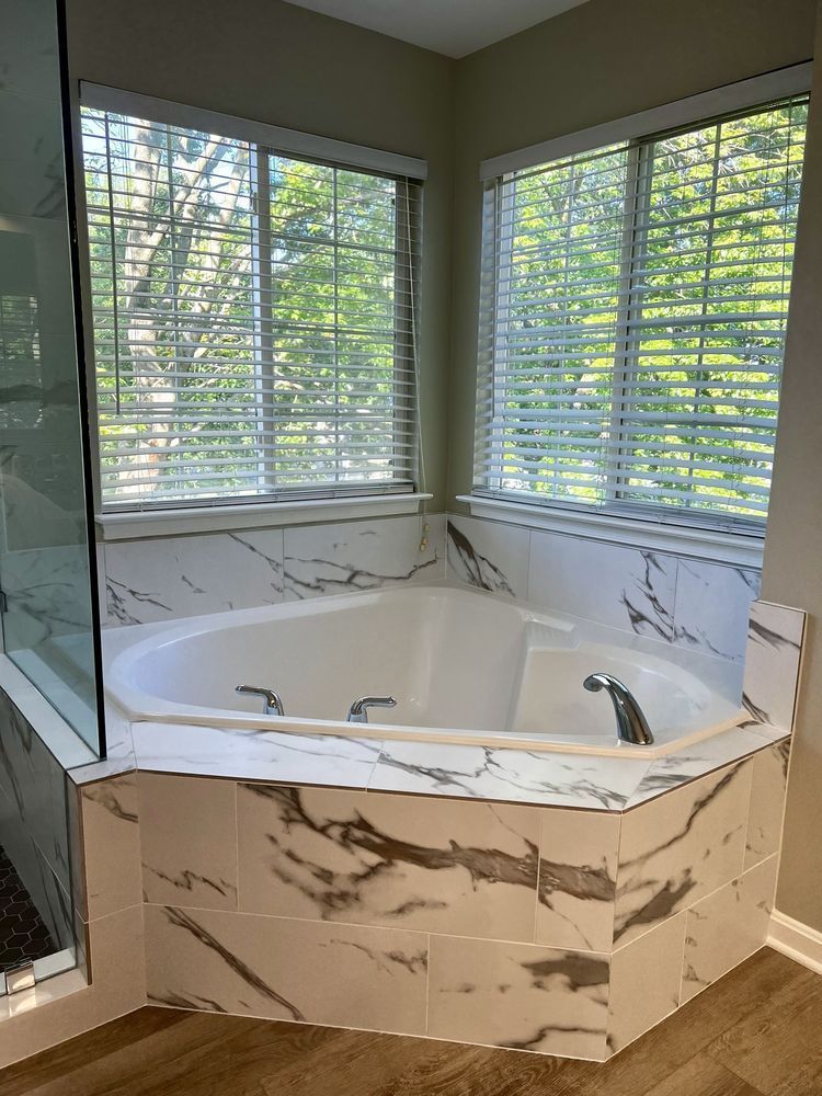 White marble-tiled jacuzzi tub in a bathroom corner, next to a glass shower. Blinds cover the windows.