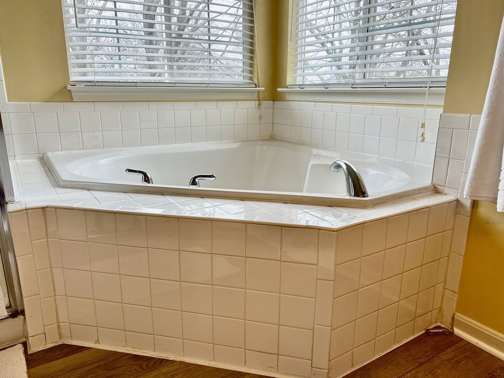Corner bathtub surrounded by white tiled walls and a window with blinds.