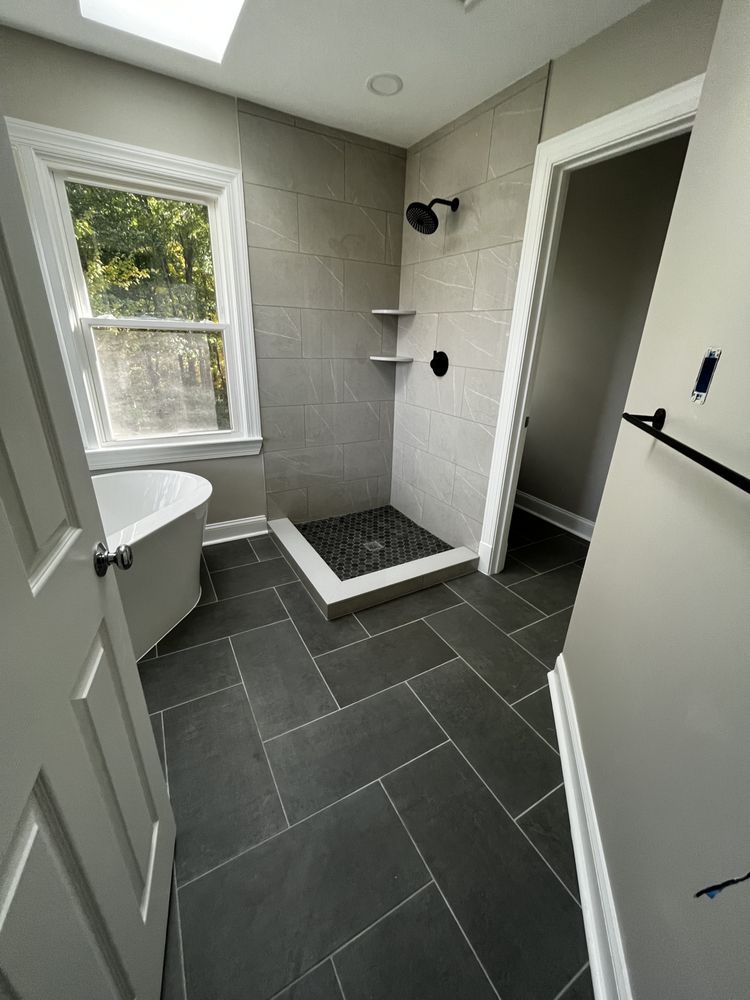 Modern bathroom with gray tiled walls and dark tile floors. Includes a shower, tub, and window.