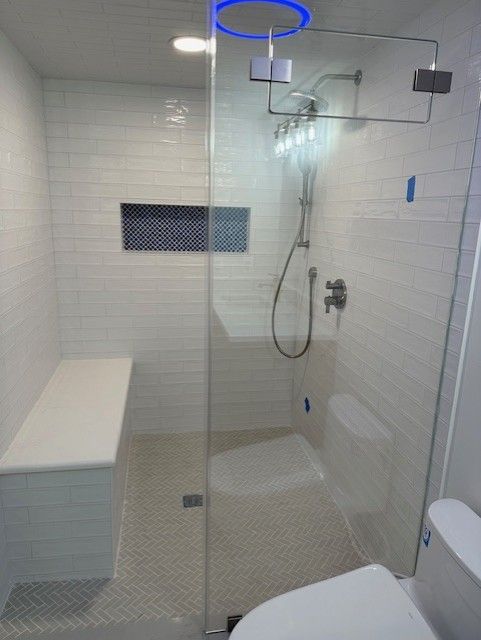 Modern white tiled shower with glass door, bench, built-in shelf, and rain showerhead; next to a toilet.