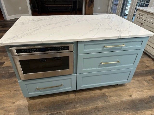 A kitchen island with a built-in microwave, drawers, and a white countertop. Blue cabinets, wood floors.