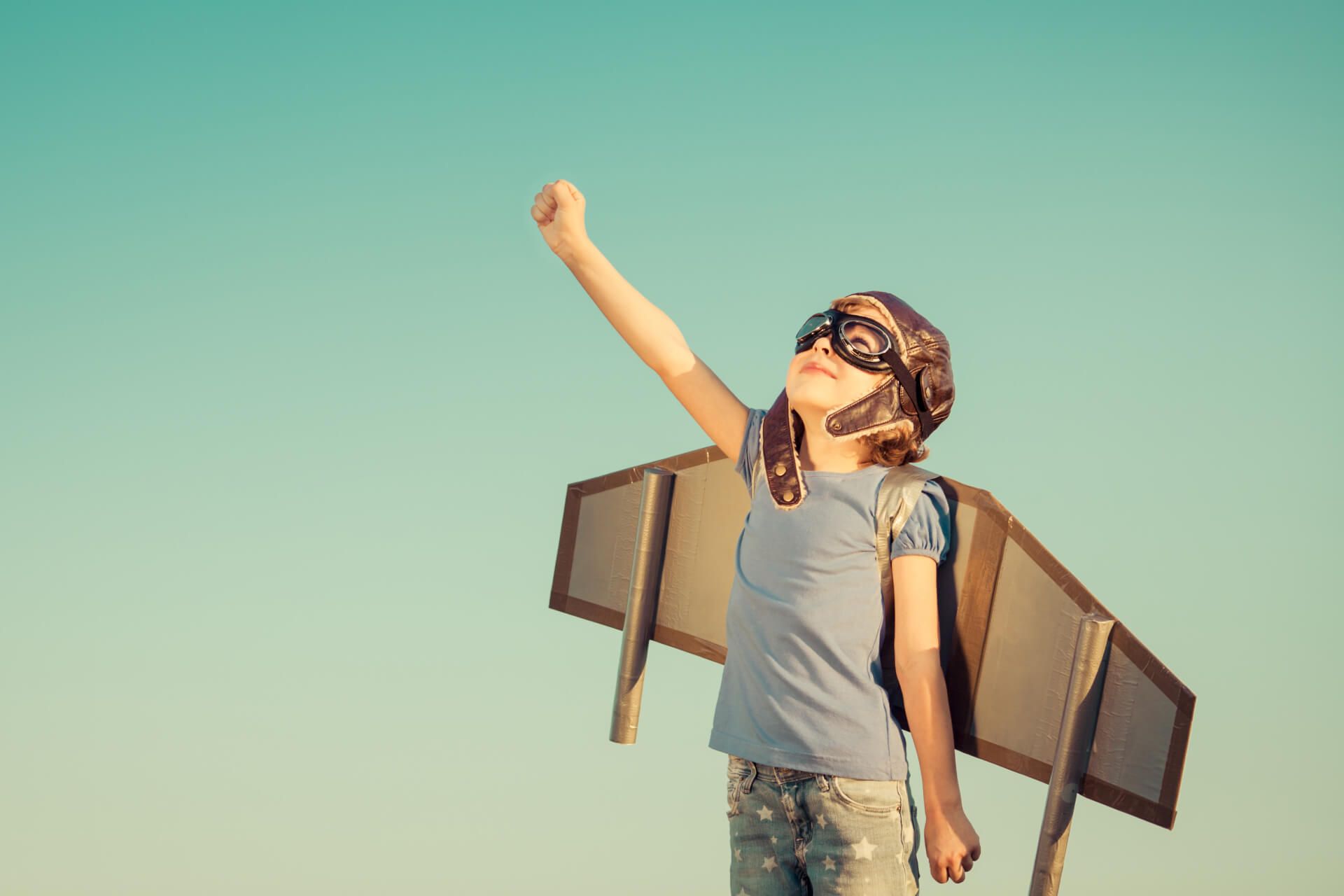 A child looks up to the sky dressed in goggles and paper wings.