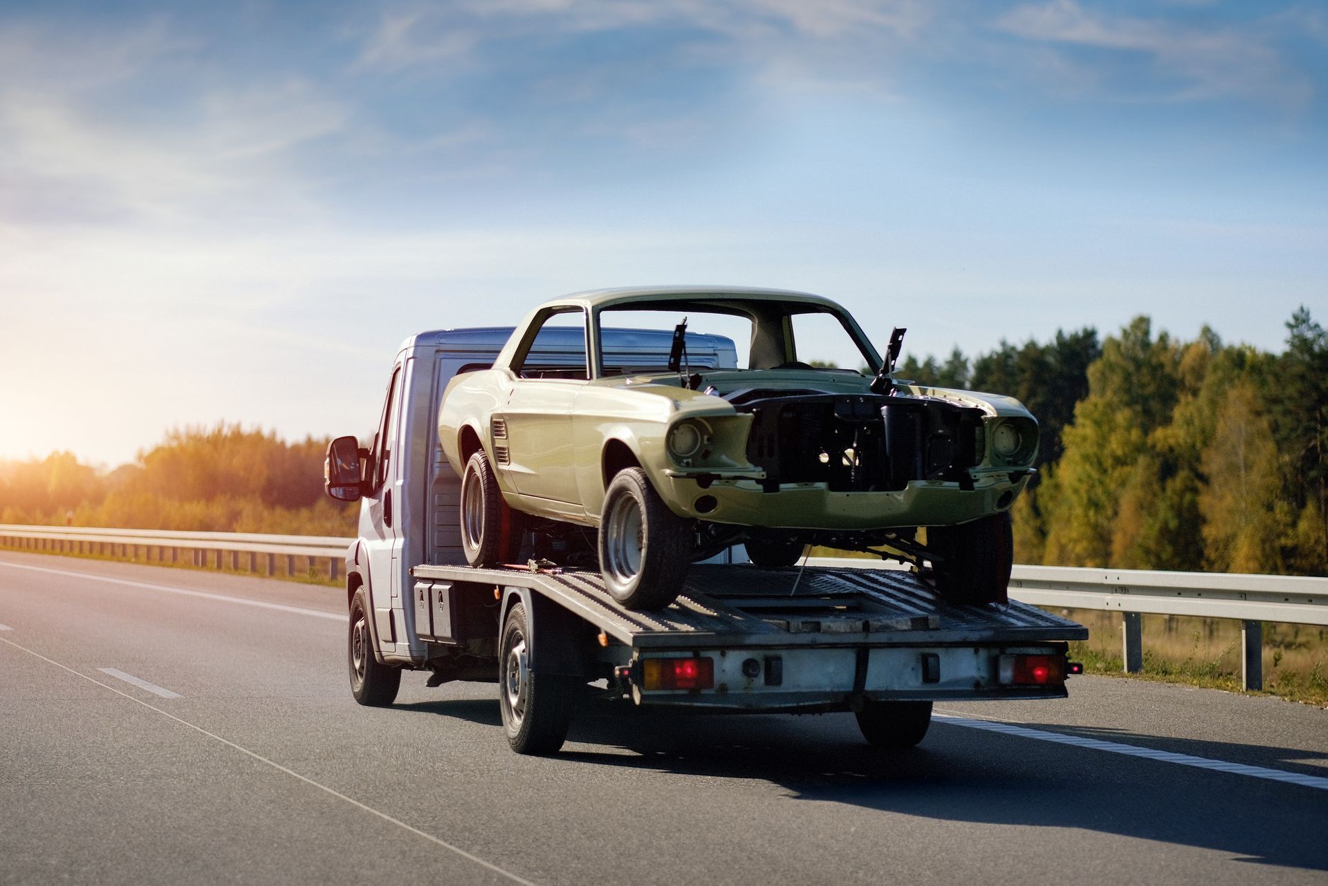 Tow truck hauling a damaged vintage car on a highway at sunset