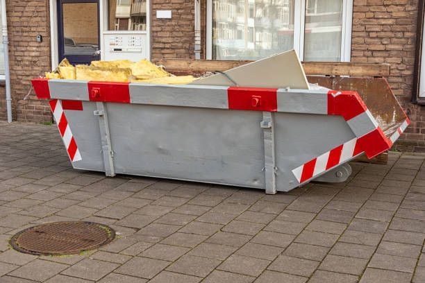 A gray skip bin filled with waste standing on a sidewalk in front of a flat.