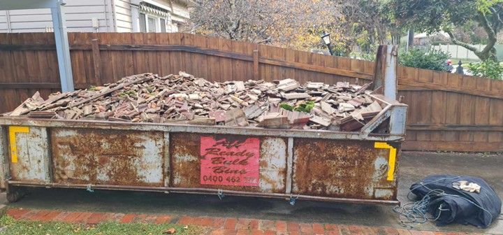 A rusty dumpster filled with broken bricks sits in a yard, next to a fence and a house.