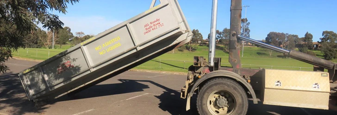 Dump truck bed raised, tilted. Truck on pavement near grassy area and trees under blue sky.