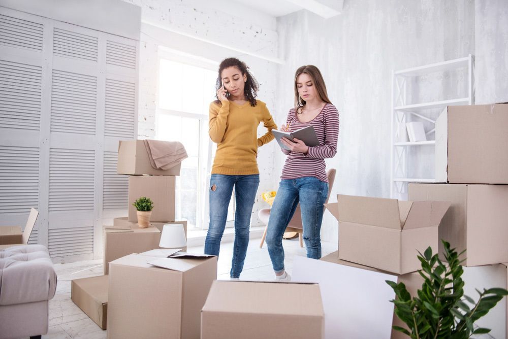 Two Women Are Standing in a Living Room Filled With Cardboard Boxes — Davis Removals & Storage in Southport, QLD