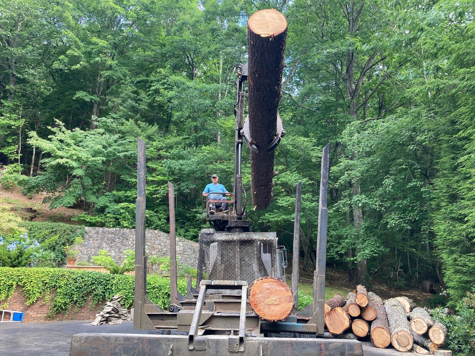 A man operates a crane lifting a large log; pile of firewood nearby; trees in the background.