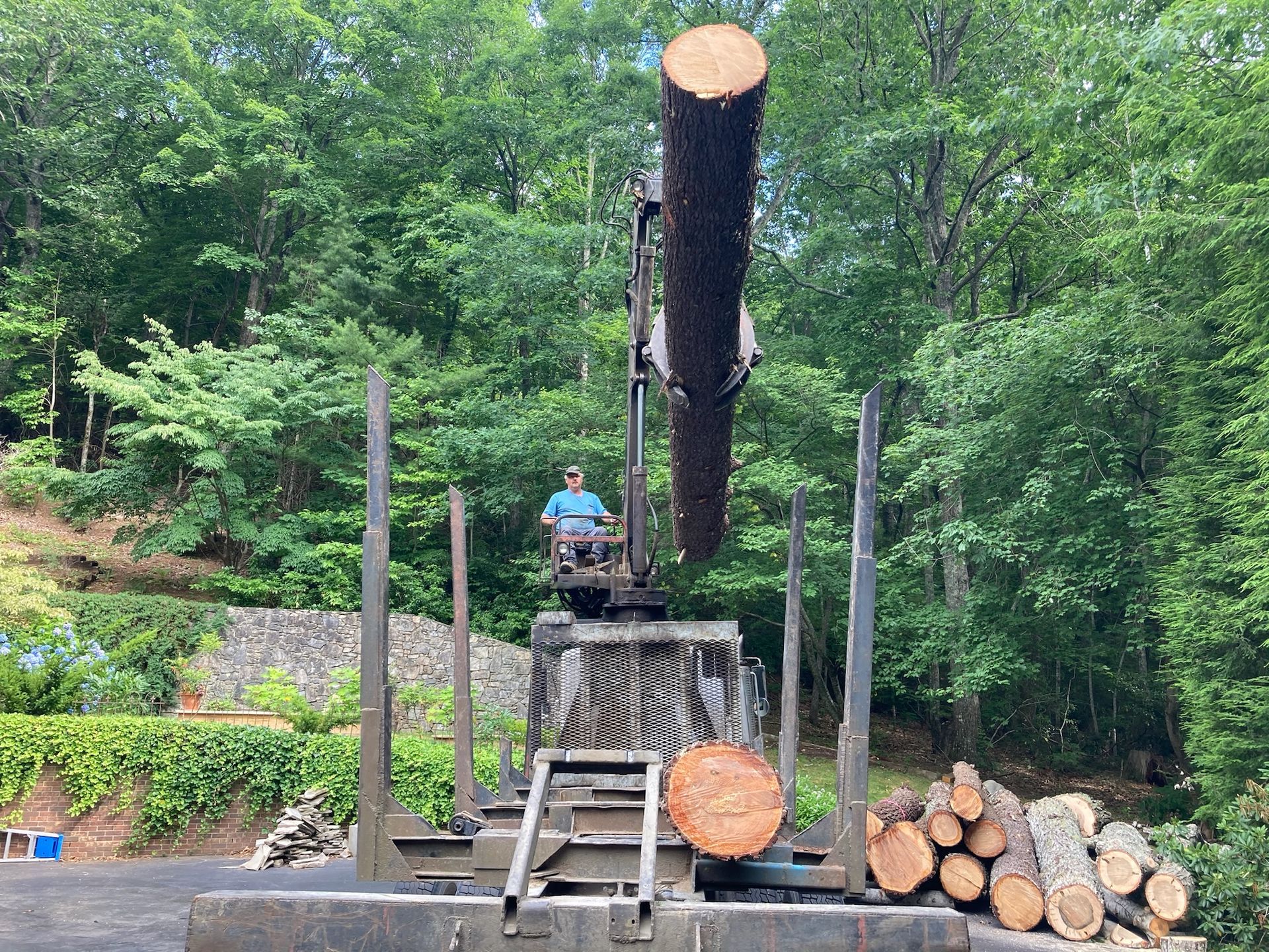 Man operating a truck-mounted crane lifting a large log; pile of cut logs on the ground, trees in background.