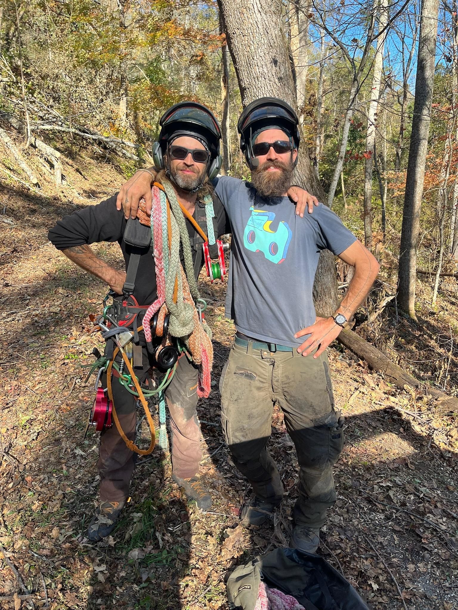 Two men in climbing gear posing in a wooded area. They wear helmets, harnesses, and have arms around each other.