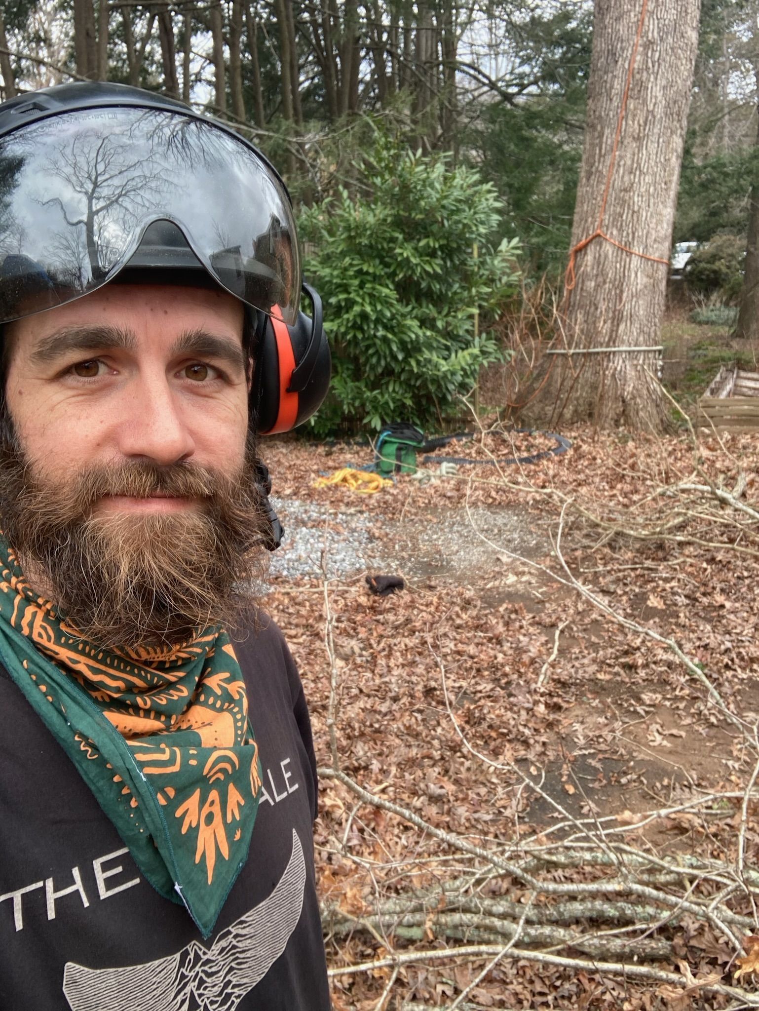 Man with beard wearing safety gear in a wooded area, surrounded by leaves and branches.
