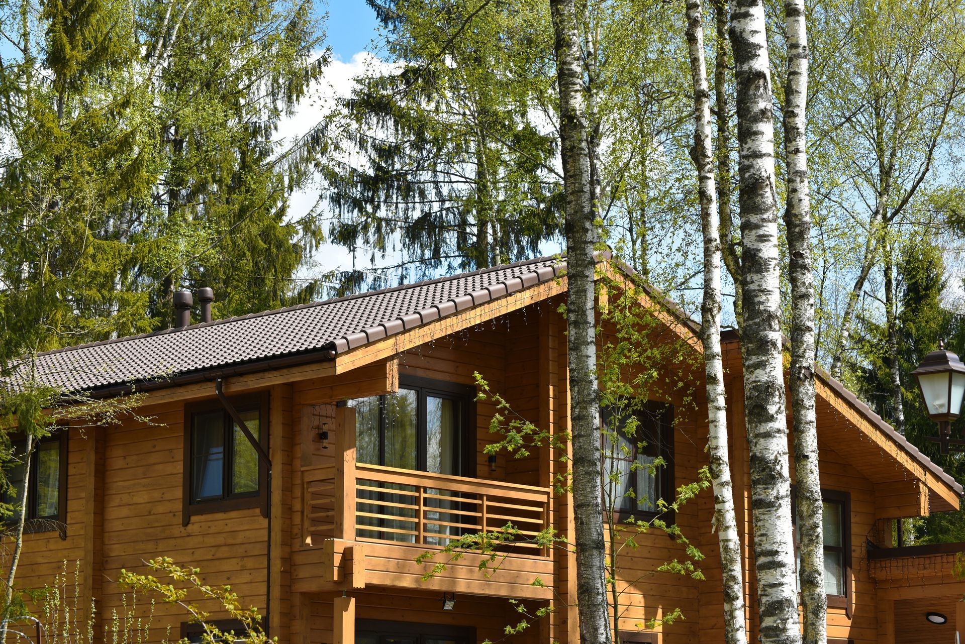 A wooden cabin nestled among birch trees with a balcony and a brown tiled roof on a sunny day.