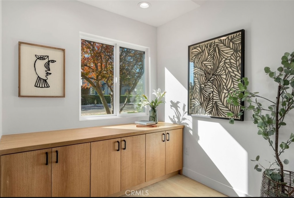 A bright room features light wood cabinets below a window, flanked by two framed artworks and a potted plant.